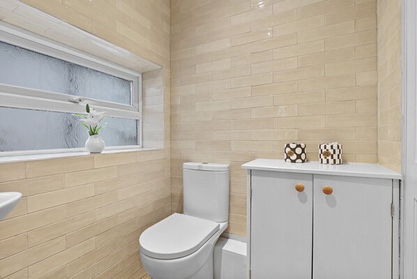 Un baño con paredes de azulejos color beige, un inodoro blanco, un lavamanos blanco y una ventana con un jarrón blanco y flores blancas.