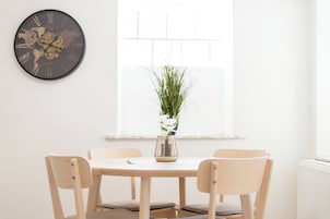 A dining area with a round table, four chairs, a world map clock, and a window with blinds.