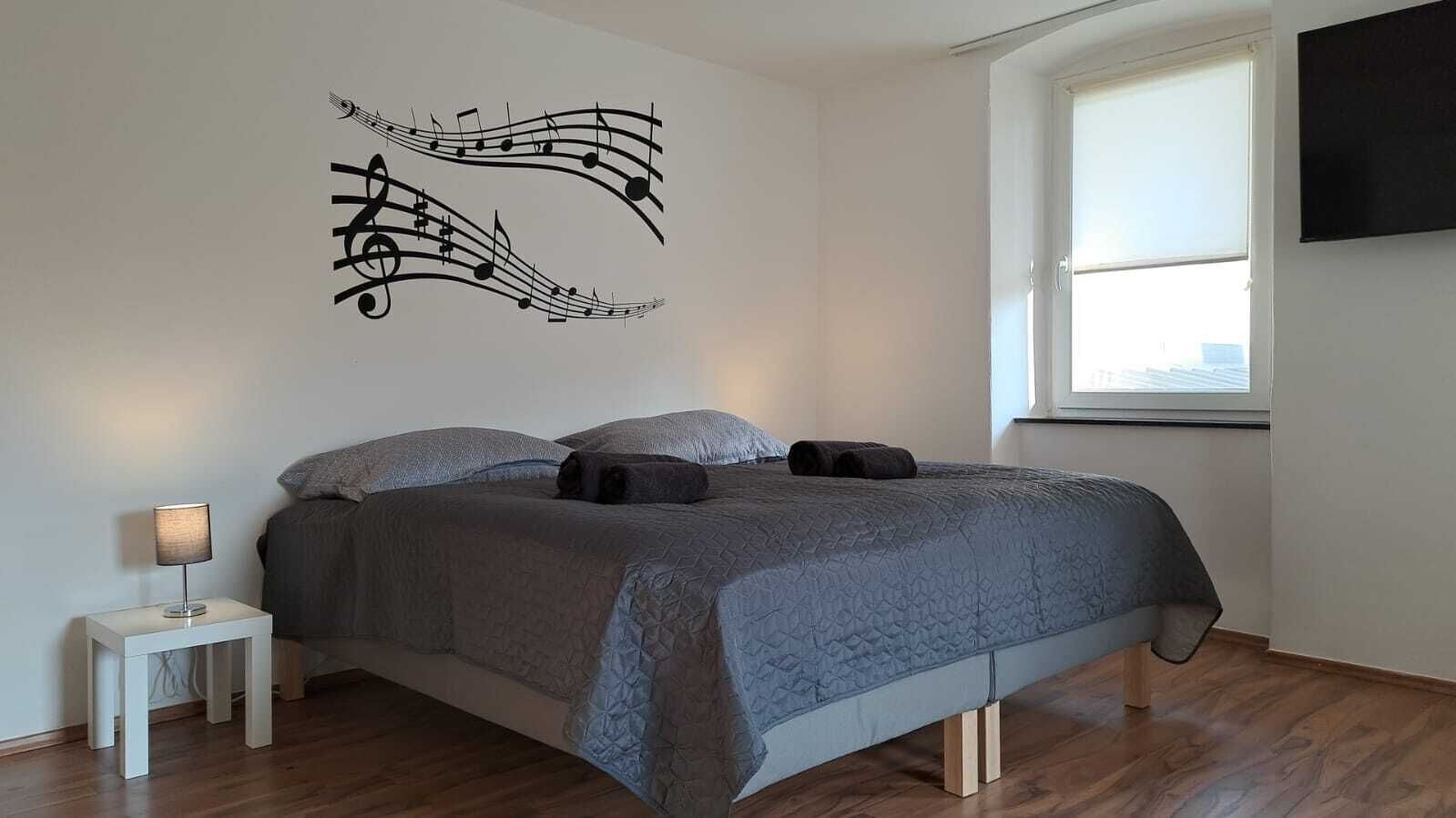 A bedroom with a bed, a nightstand, a wall art of musical notes, and a window.