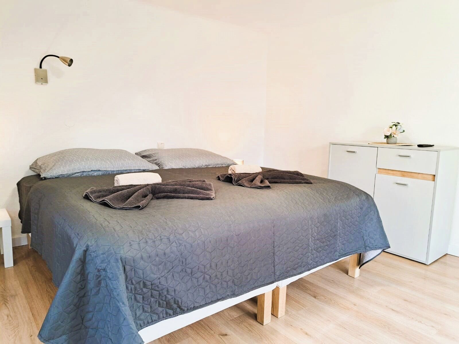 Una cama bien hecha con un edredón gris acolchado, dos almohadas estampadas y una lámpara de noche.
