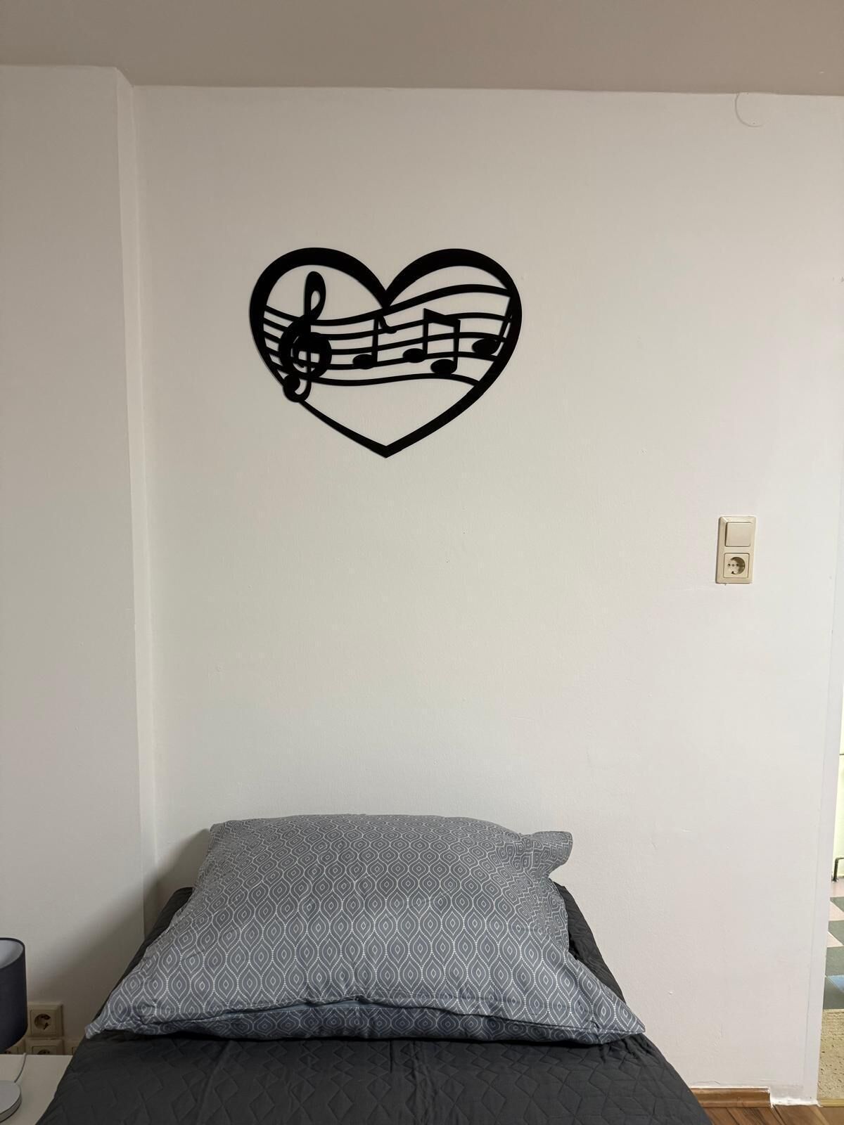 Une chambre avec un lit, un mur orné d’une décoration en forme de cœur représentant des notes de musique, et une prise de courant.