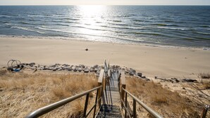 Une plage avec un escalier en bois descendant jusqu’au rivage, entourée d’herbes sèches et de rochers.