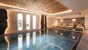 A modern indoor pool area with a wooden ceiling, large windows, and a seating area with cushioned chairs. - DISTINCTIVE LUXURY DOLOMITI APARTMENTS TERRE71 (Marebbe)