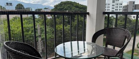 A balcony with two wicker chairs and a glass table, overlooking a cityscape with buildings and greenery.