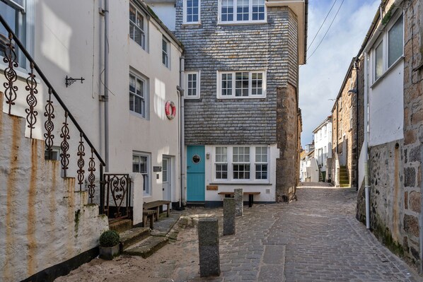 A narrow cobblestone street flanked by historic stone buildings with white and blue doors, a bench, and a metal staircase.