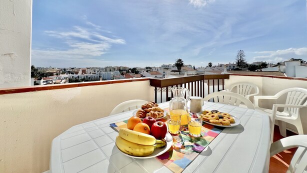 En balkon med et bord dækket til morgenmad, inklusive frugt, bagværk og drikkevarer, med udsigt over byens skyline.