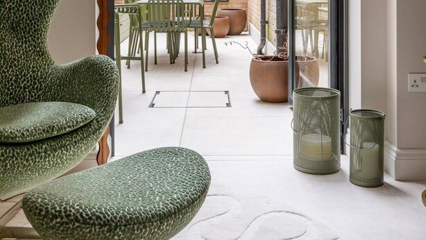 A modern interior with a green patterned chair, a table, and a lamp. There is a view of an outdoor area with a table and chairs, a brick wall, and greenery.