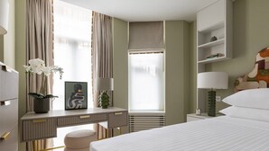 A modern hotel room with a bed, bedside tables, a desk, a chair, and a window with curtains.