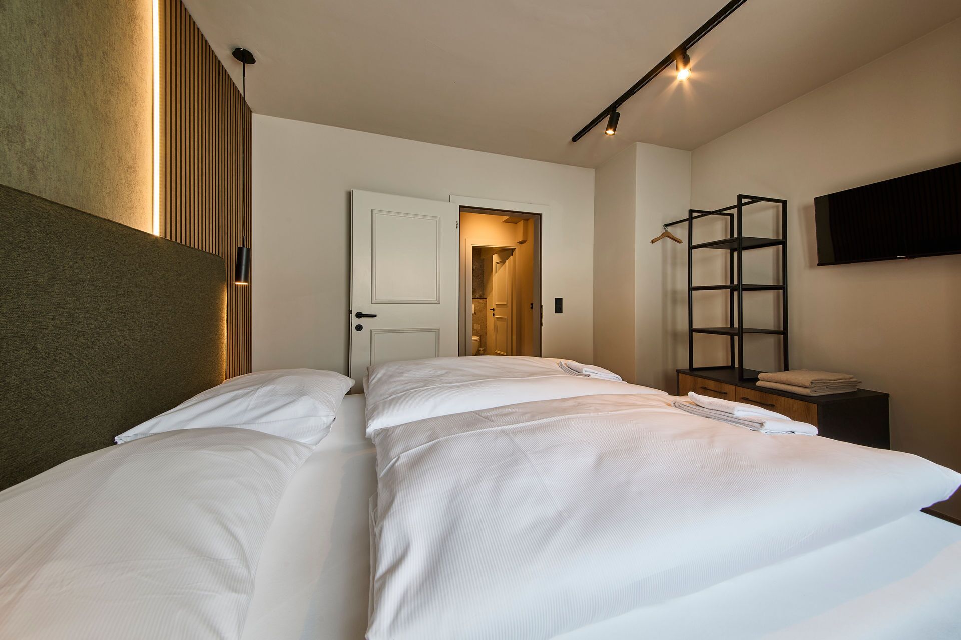 A hotel room with two beds, a wooden headboard, a TV mounted on the wall, and a shelf with towels.