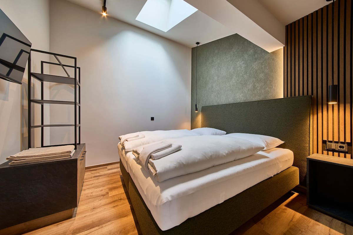 Une chambre moderne avec un puits de lumière, un lit sur lequel sont soigneusement pliées des serviettes, une table de chevet en bois et un luminaire fixé au mur.