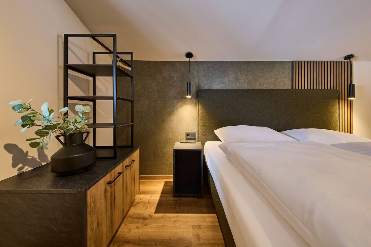 Une chambre moderne avec une commode en bois, une étagère en métal noir, une table de chevet avec une lampe et un lit recouvert de linge de maison blanc.