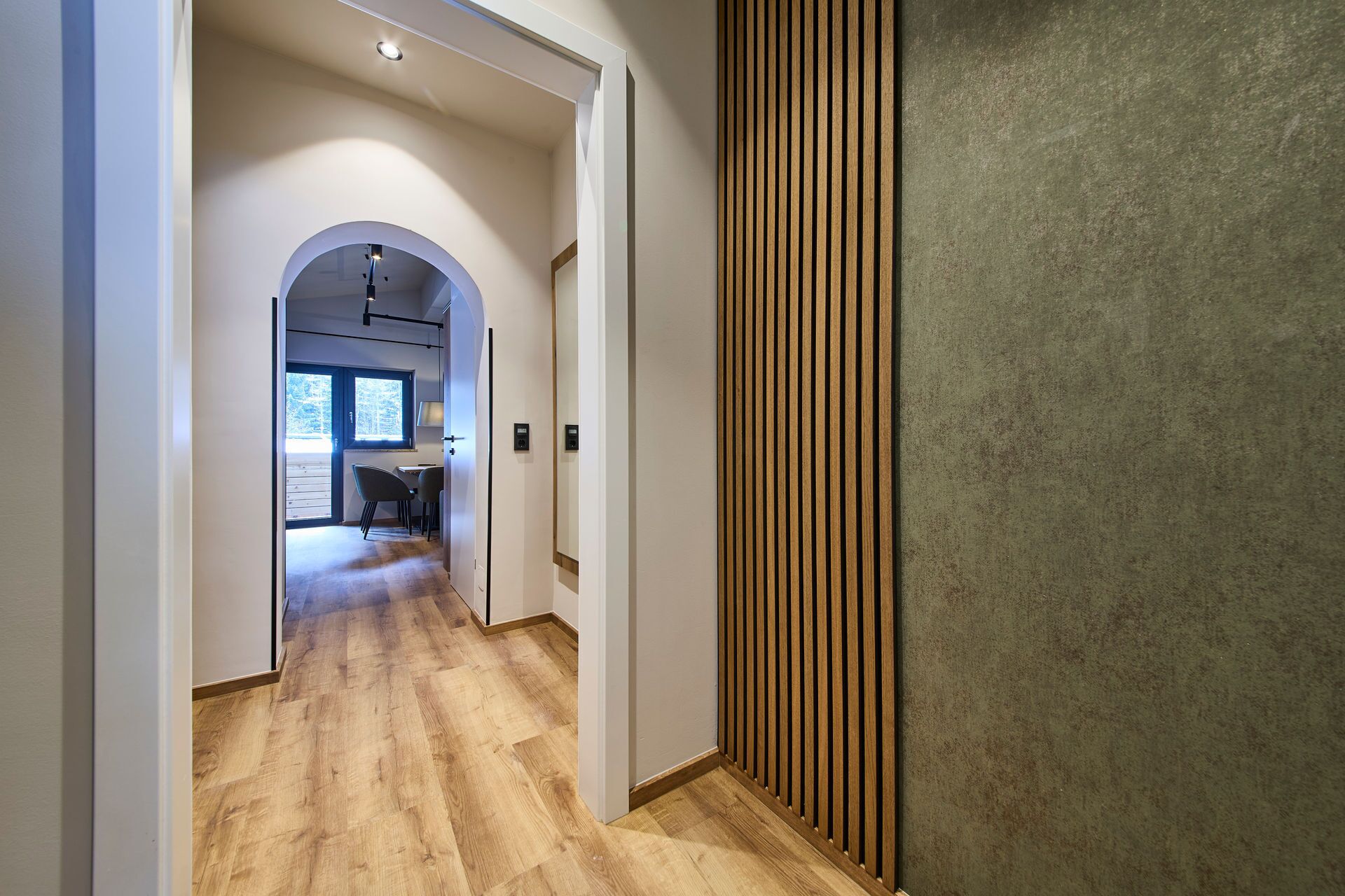 A hallway with wooden flooring, a modern interior design, and a view into a room with a desk and chairs.