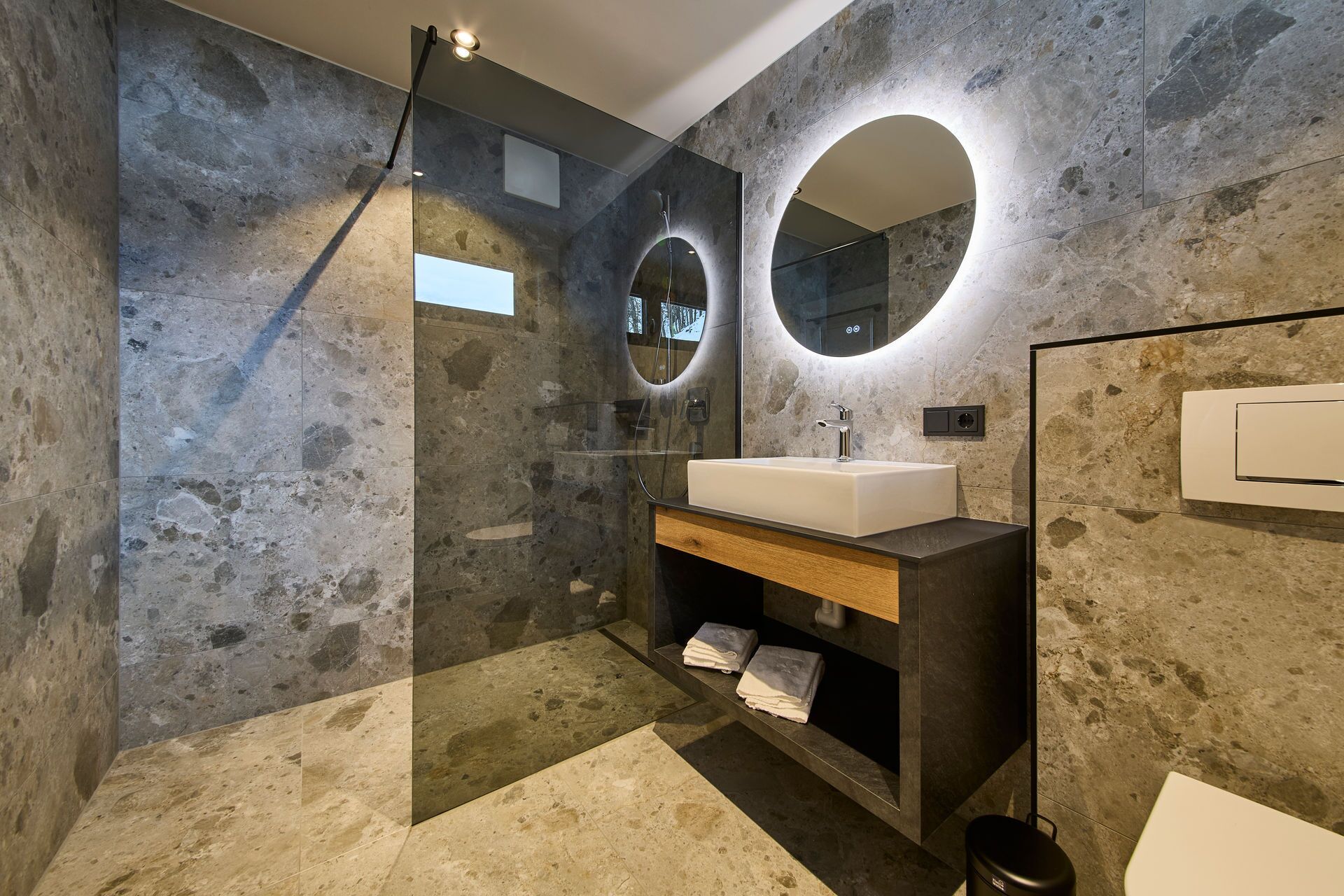 A modern bathroom with a stone wall, a glass shower enclosure, a sink with a wooden vanity, and a round mirror.