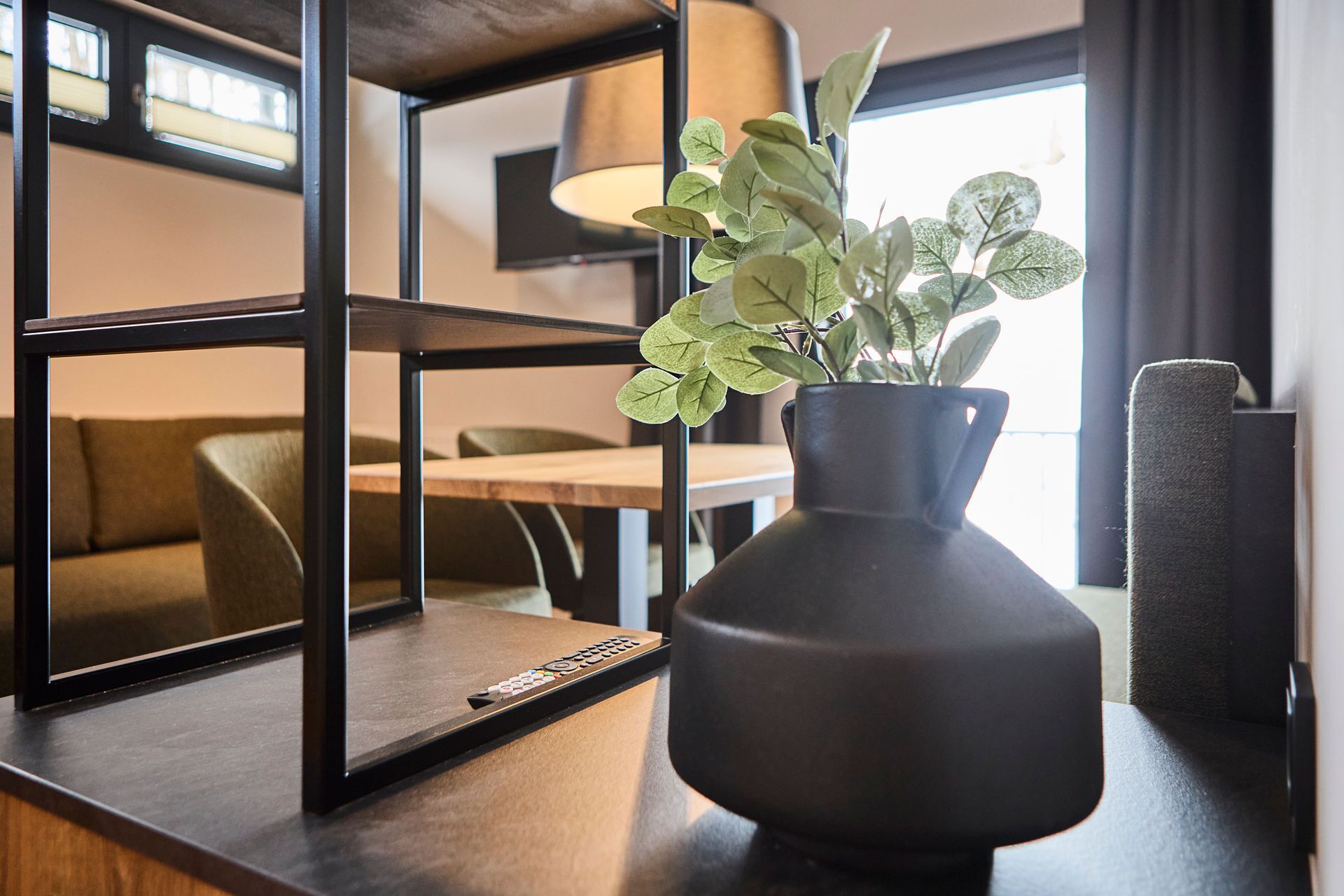 Un interior moderno con una mesa de madera oscura, un jarrón negro con hojas verdes y un espejo grande que refleja una habitación con un sofá y una lámpara.