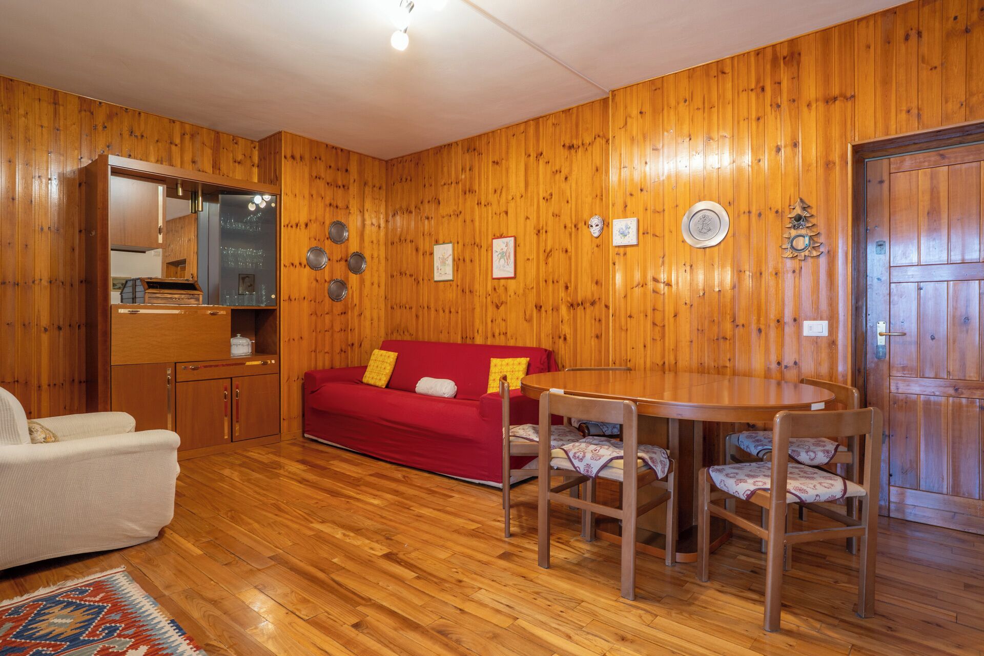 A cozy room with wooden walls, a red sofa, a white armchair, a wooden dining table with four chairs, and a small kitchen area with a mirror and cabinets.