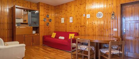 A cozy room with wooden walls, a red sofa, a white armchair, a wooden dining table with four chairs, and a small kitchen area with a mirror and cabinets.