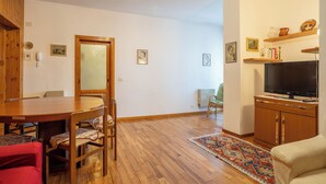A room with a wooden dining table and chairs, a television on a cabinet, and a patterned rug on the floor. - Maggio - appartamento con piscina (Cremeno)