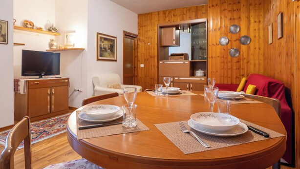 A dining area with a wooden table set for four, surrounded by chairs. A television is mounted on the wall to the left. - Maggio - appartamento con piscina (Cremeno)