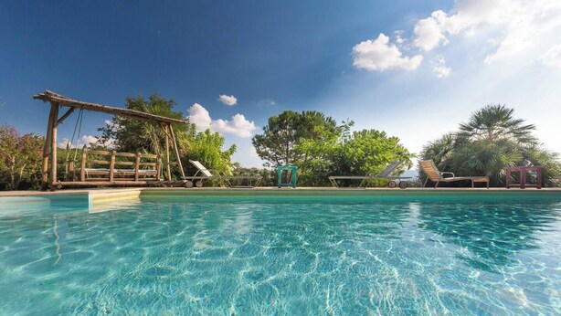 A clear pool with a wooden deck and lounge chairs, surrounded by trees and a clear sky.