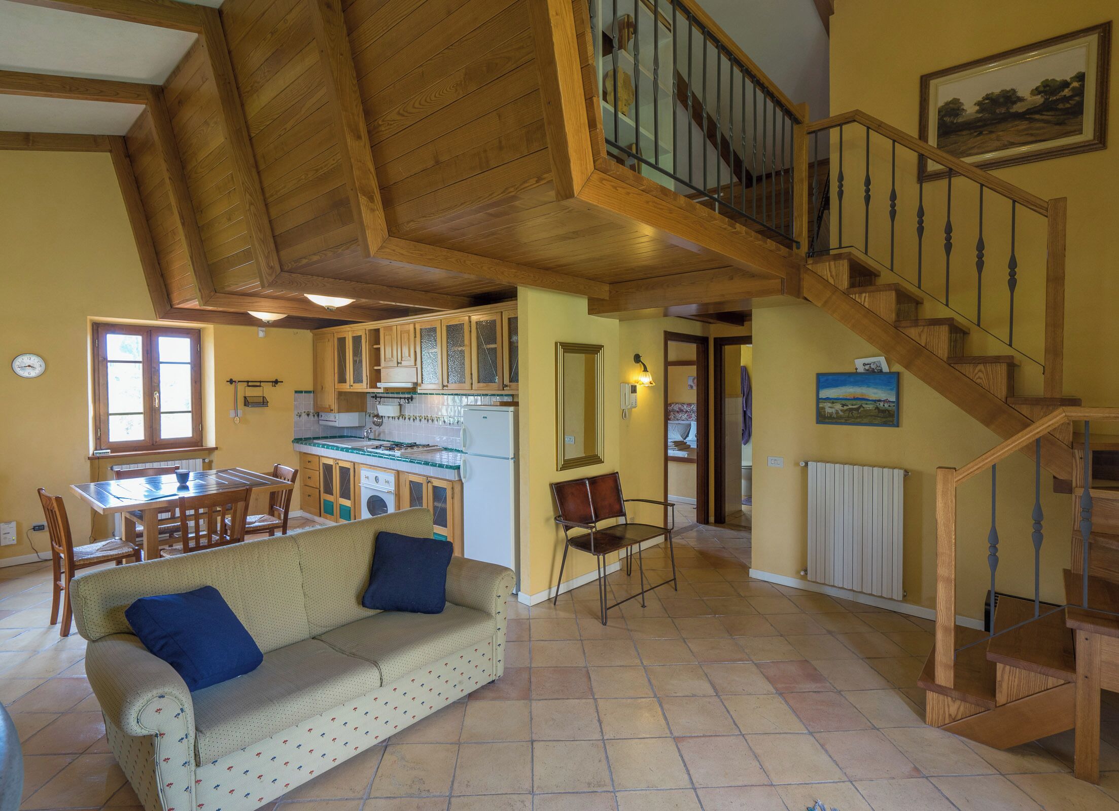 A spacious living area with a wooden staircase, a dining table with chairs, a sofa with cushions, and a kitchen area with wooden cabinets and a refrigerator.