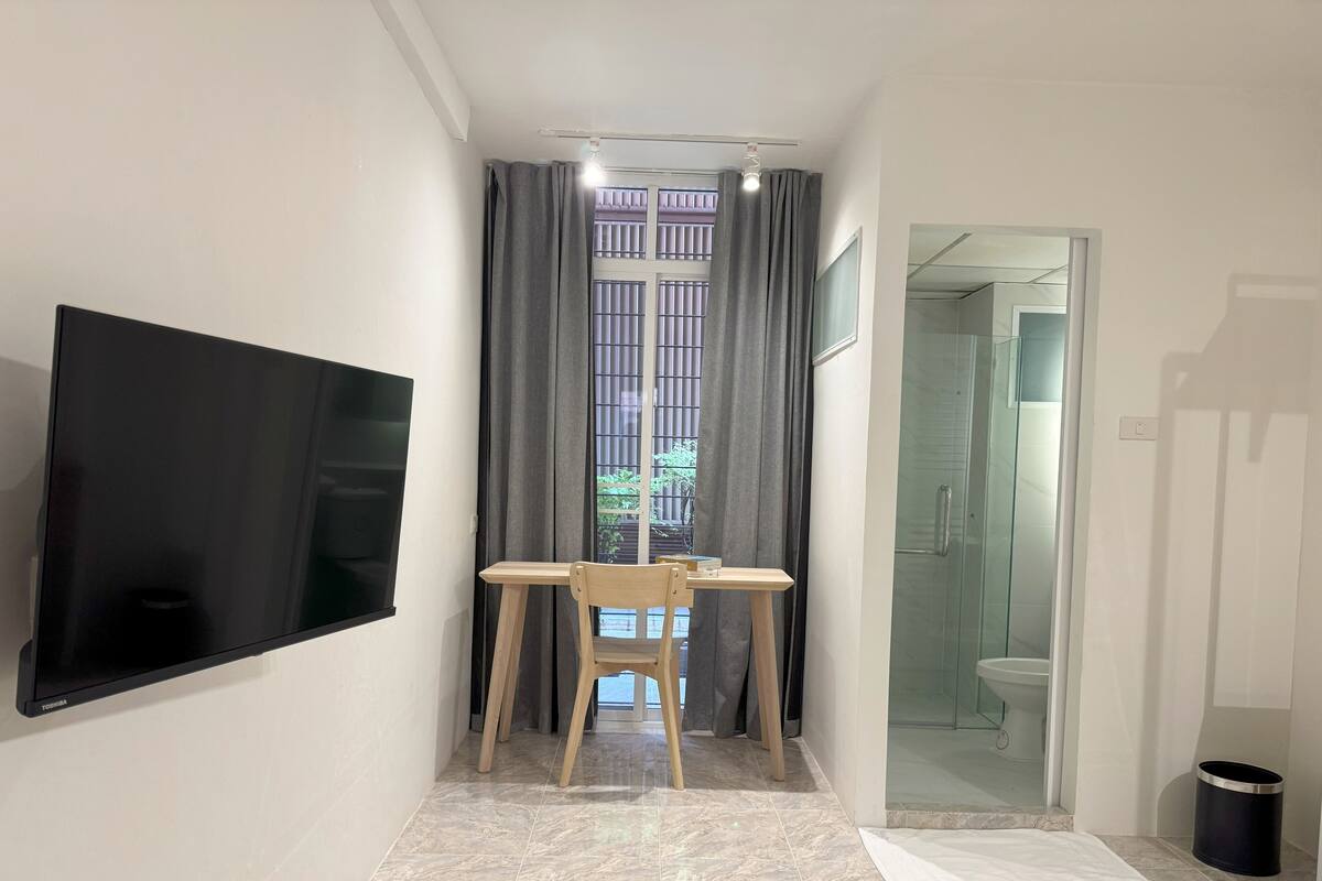 A small, modern room with a flat-screen TV mounted on the wall, a wooden table with a chair, a window with curtains, and a glass shower area.