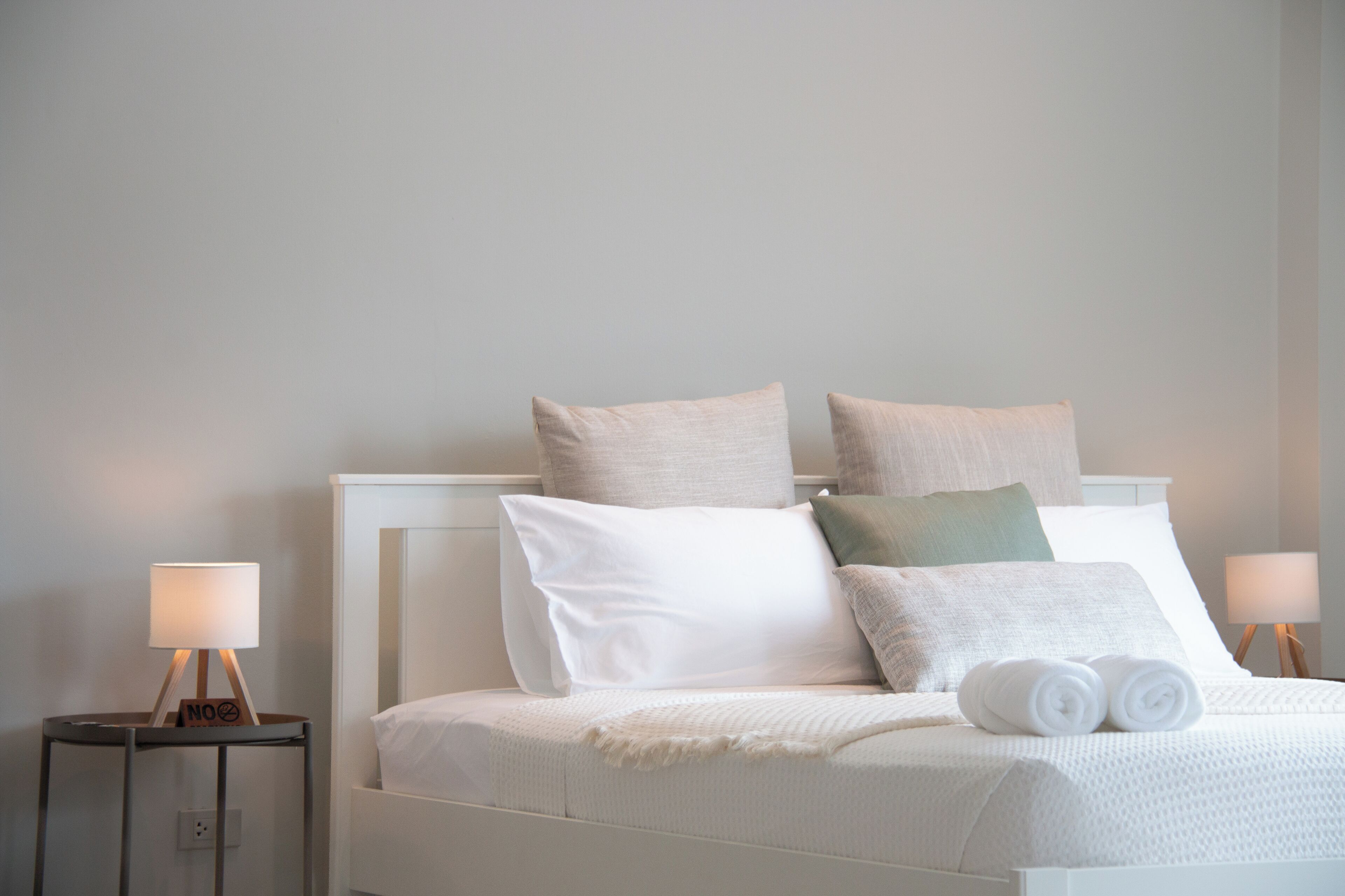 A neatly made bed with white bedding and multiple pillows, two bedside tables with lamps, and a plain wall in the background.