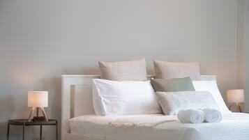 A neatly made bed with white bedding and multiple pillows, two bedside tables with lamps, and a plain wall in the background.