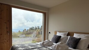 Una camera da letto con un'ampia finestra, un letto con lenzuola bianche, una coperta grigia a motivi e una testiera in legno.