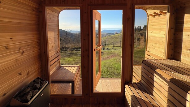 Una baita di legno con una panca e un tavolino, una porta che dà all'esterno e vista su un paesaggio verde e delle colline.
