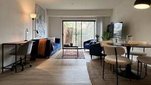 A modern living room with a sofa, coffee table, and dining area. There is a large window with a view of greenery outside. - Serenity Retreat in Santa Monica (Santa Monica)