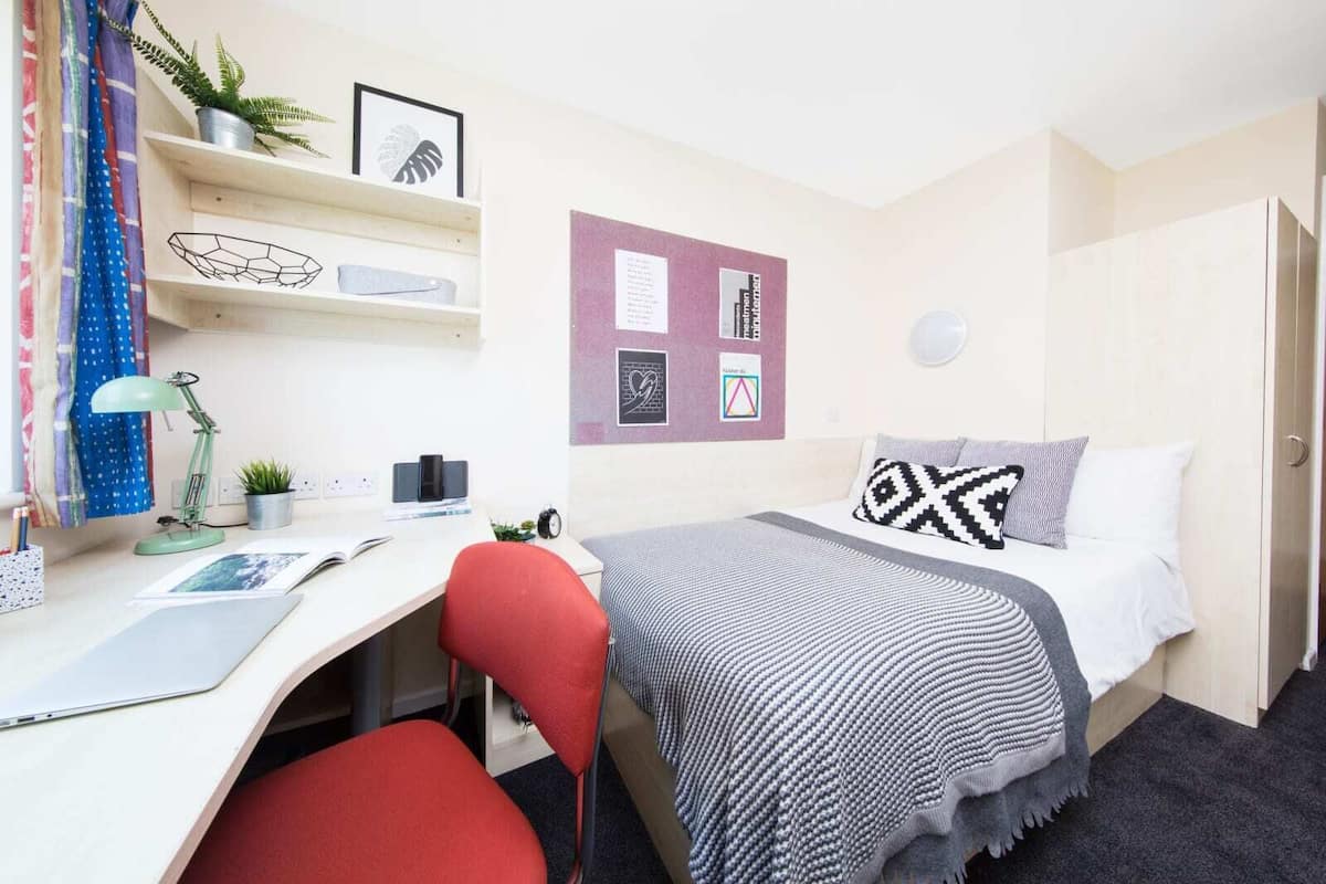 A compact bedroom with a bed, a desk, a chair, a wall-mounted bulletin board, and a potted plant.