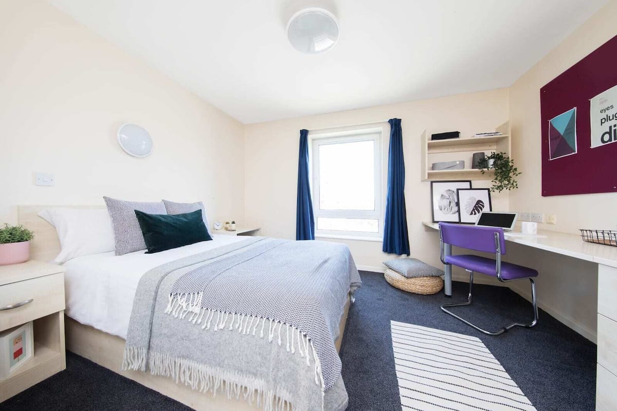 A modern bedroom with a bed, bedside table, desk, chair, and a window with curtains.
