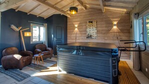 Une pièce avec un plafond en bois, un grand jacuzzi noir, deux canapés en cuir marron, une petite table en bois et une fenêtre avec des rideaux.