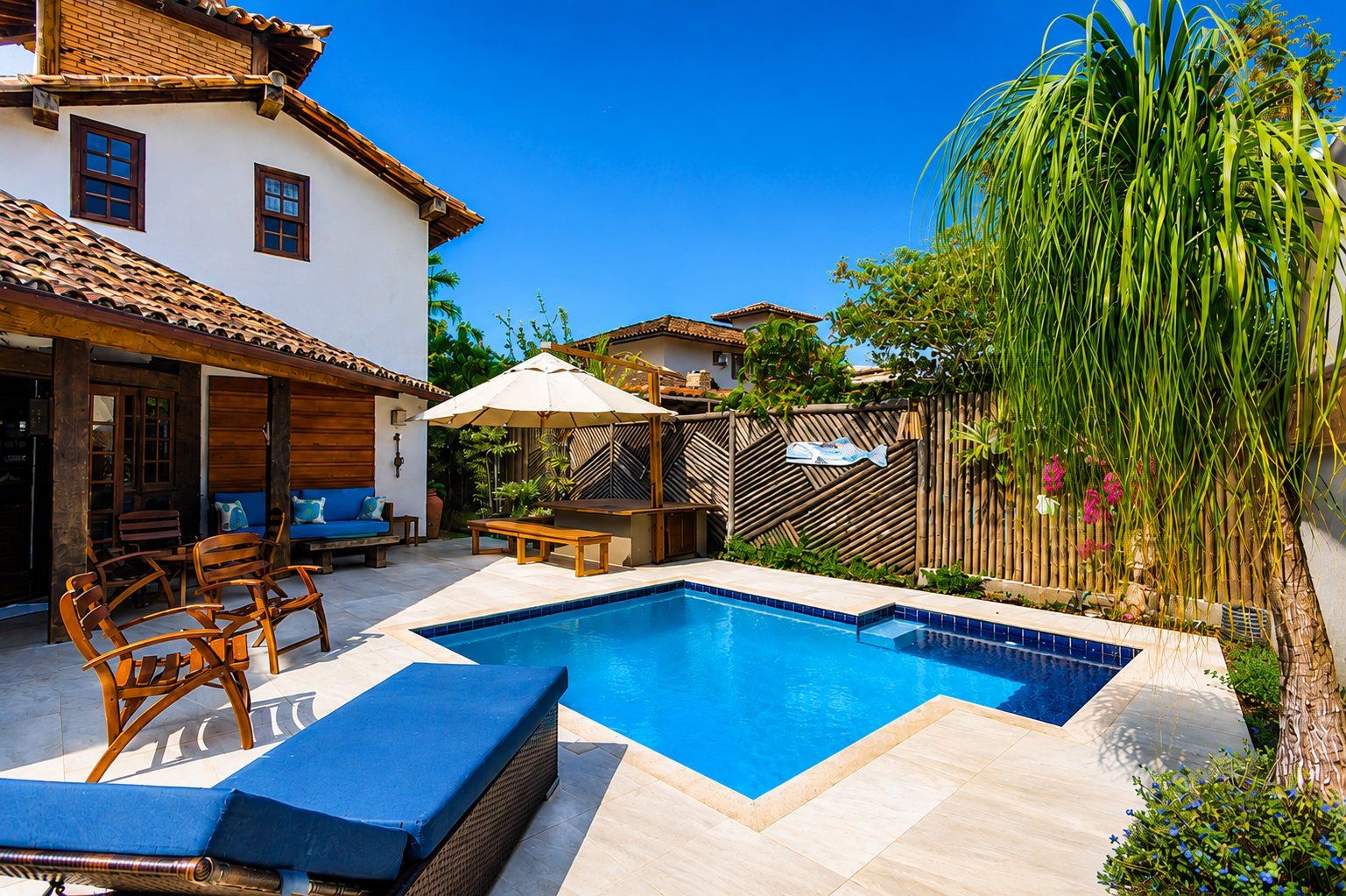 A swimming pool with a tiled deck, wooden furniture, and a bamboo fence.
