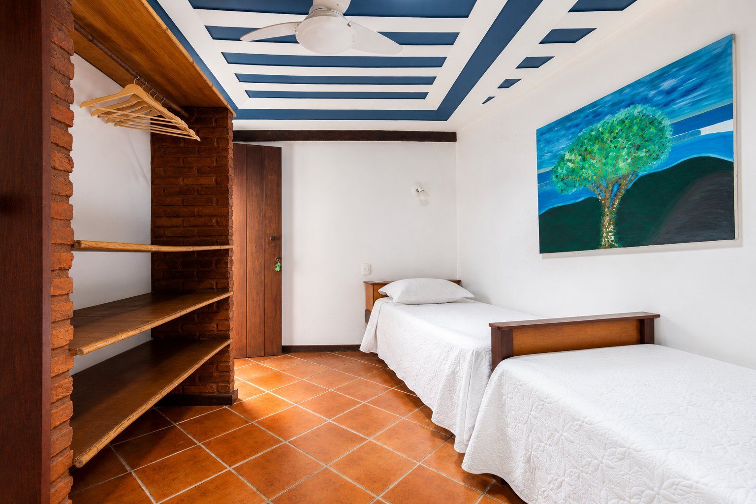A bedroom with two beds, a wooden shelf unit, a ceiling fan, and a painting of a tree.