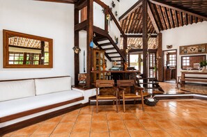 A spacious room with a high ceiling, wooden beams, a tiled floor, a white sofa, and a dining area with chairs and a table.