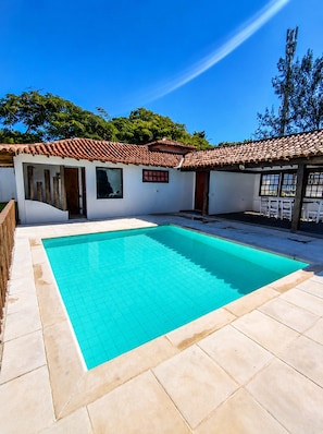 A swimming pool with tiled edges, a covered patio area, and a building with a tiled roof. - Bz34 Casa 150m da praia Piscina e Sauna (Buzios)