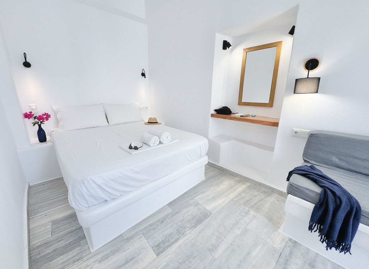 Una habitación de hotel moderna con una cama grande, una mesita de noche con un florero, una pequeña mesa con un sombrero, un espejo y un sofá con una manta.