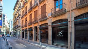Une rue bordée d’un immeuble doté d’une grande façade en verre sur laquelle figure un logo, ainsi que d’un trottoir piétonnier et de bornes le long du bord de la chaussée.