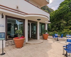 Entrada de hotel con puerta de cristal, plantas en macetas y mesas al aire libre.