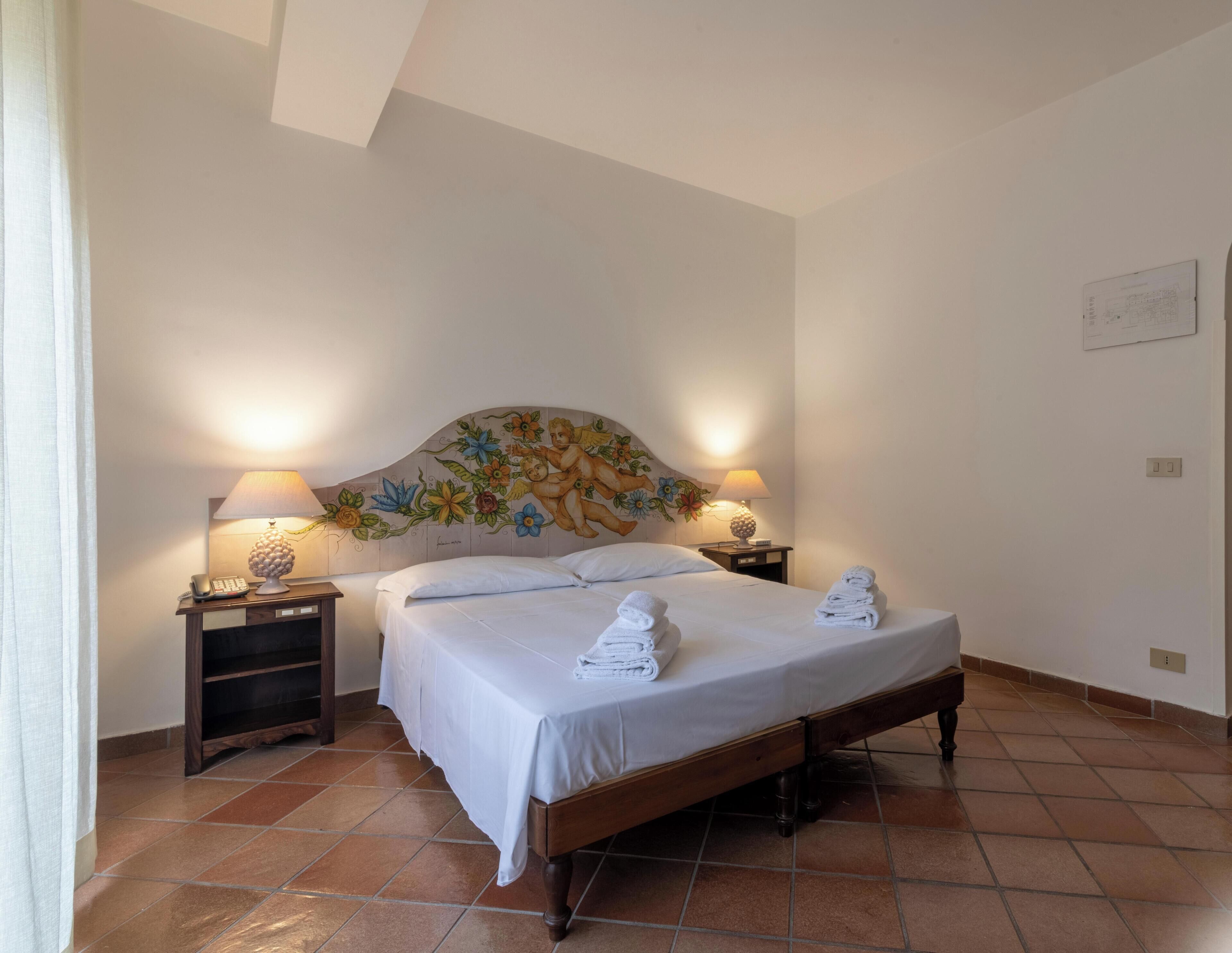 A hotel room with a bed, two bedside tables with lamps, a headboard with a floral mural, and a tiled floor.
