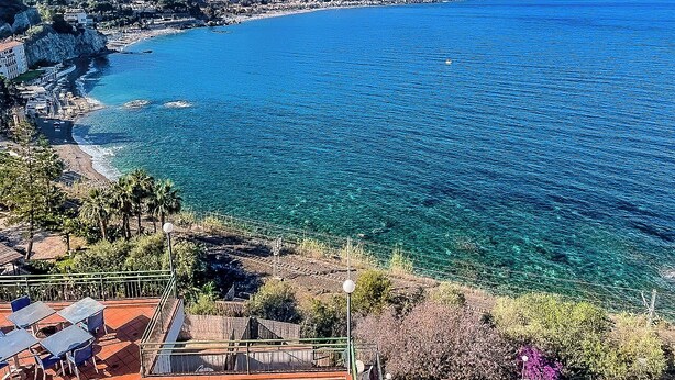 A coastal view with clear blue waters, a beach, and buildings along the shore. - Hotiday Taormina Mare (Taormina)