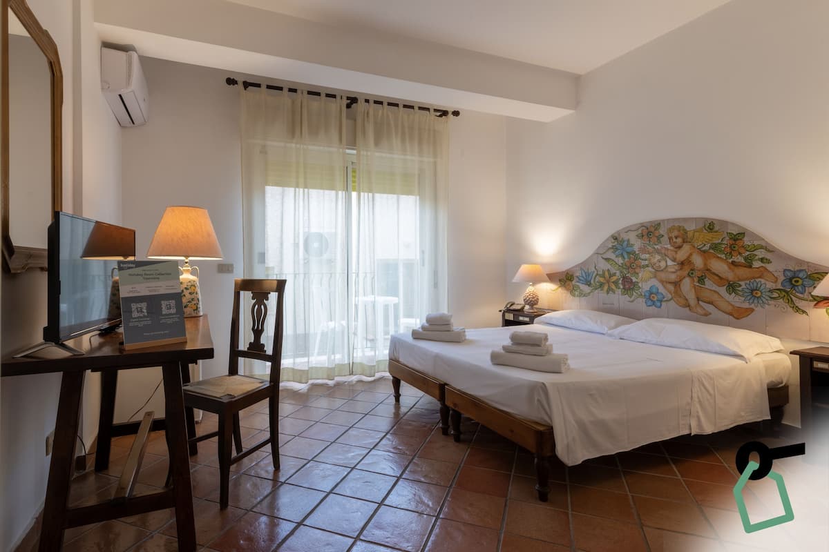 A hotel room with a bed, a desk with a computer and lamp, a chair, a window with curtains, and a decorative headboard.