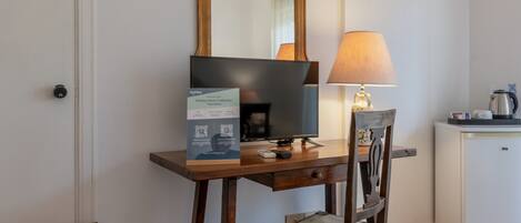 A hotel room with a wooden desk, a chair, a television, a lamp, a small refrigerator, and an air conditioning unit.