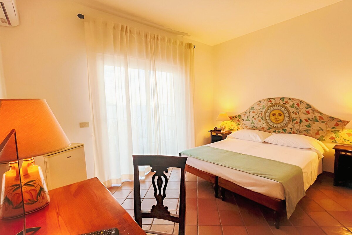 A hotel room with a bed, two bedside lamps, a desk with a lamp, a chair, and a window with curtains.