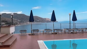 A poolside area with lounge chairs and umbrellas, a tiled deck, and a view of the sea and mountains. - Hotiday Taormina Mare (Taormina)