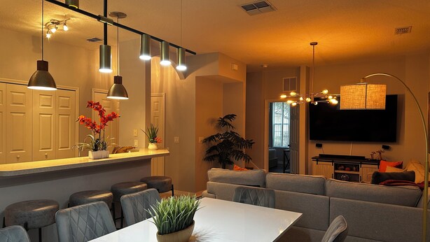 A modern living room with a white dining table, grey sofas, and a flat-screen TV mounted on the wall. - Cozy townhouse close to Disney, Universal parks, restaurant with prkg free (Kissimmee)