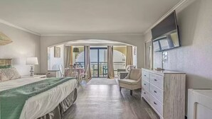 A hotel room with a bed, a desk, a chair, a dresser, and a TV. There is a balcony with curtains and a view of the outside.