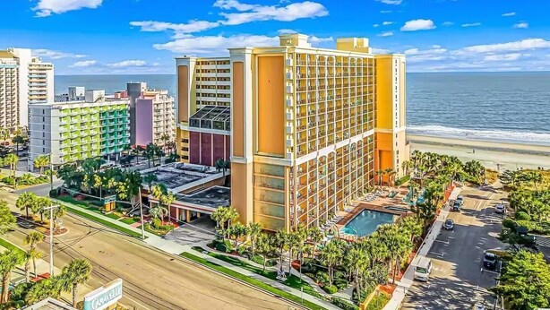 A hotel with a pool, located near a beach with palm trees and a clear blue sky.