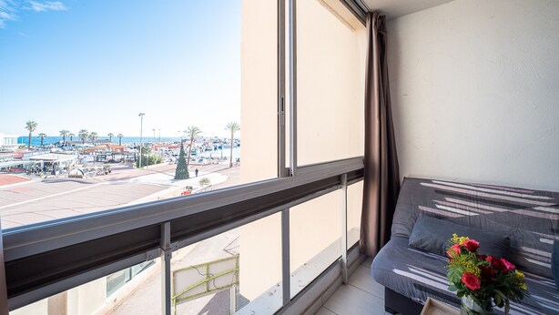 A balcony with a view of a busy street and palm trees, a sofa with a vase of flowers, and a window with curtains. - Clématite - Vue mer, plage à 100m, proche Espagne (Saint-Cyprien)